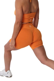 Woman in orange scrunch bum shorts and tank top, close-up of shoe logo on fabric, gym wear for ultimate curve control.